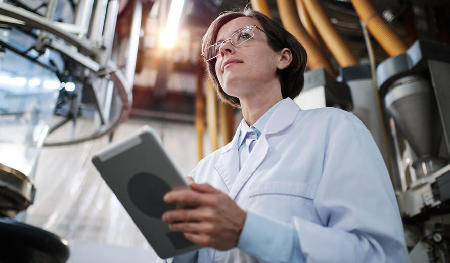 Portrait of female factory worker wearing glasses and white robe, using tablet for quality control and logistic purposes at polymer plastic manufacturing, standing between shelves with polymer rolls