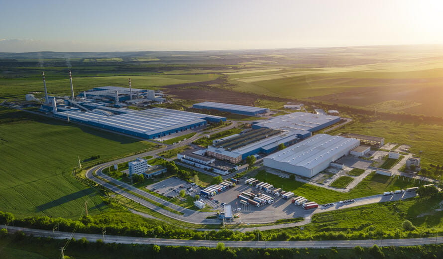 Drone view over big factory among vivid greenery on a bright summer day. Sustainability and coexistance of nature and industry.