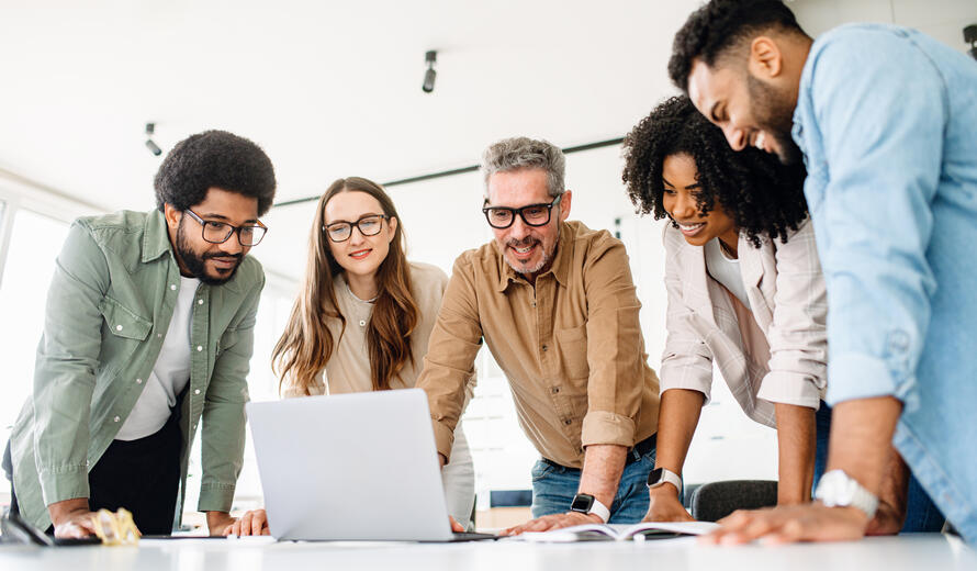 A group of diverse professionals is standing around a laptop, highlighting a moment of team collaboration and digital strategy planning, solving project tasks, or analyzing the result