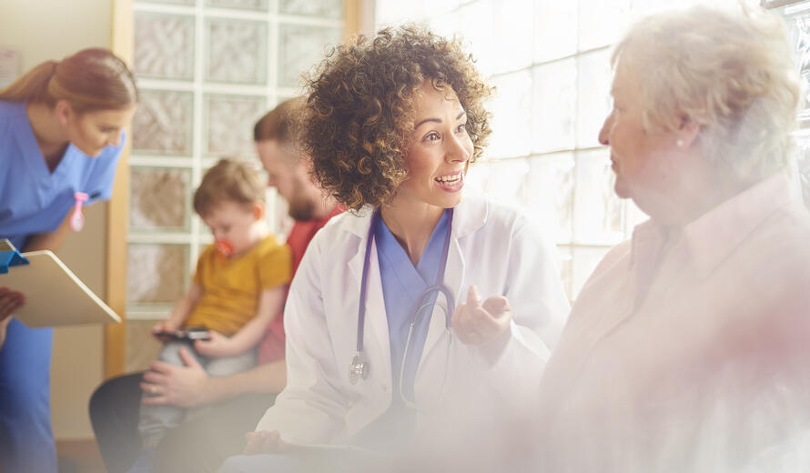 A female nurse sits in doctor’s waiting room with senior female patient taking notes and updating her medical records, another patient checks his details are correct with another nurse in the background