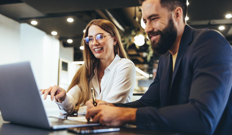 Cheerful businesspeople using a laptop in an office. Happy young entrepreneurs smiling while working together in a modern workspace. Two young businesspeople sitting together at a table.